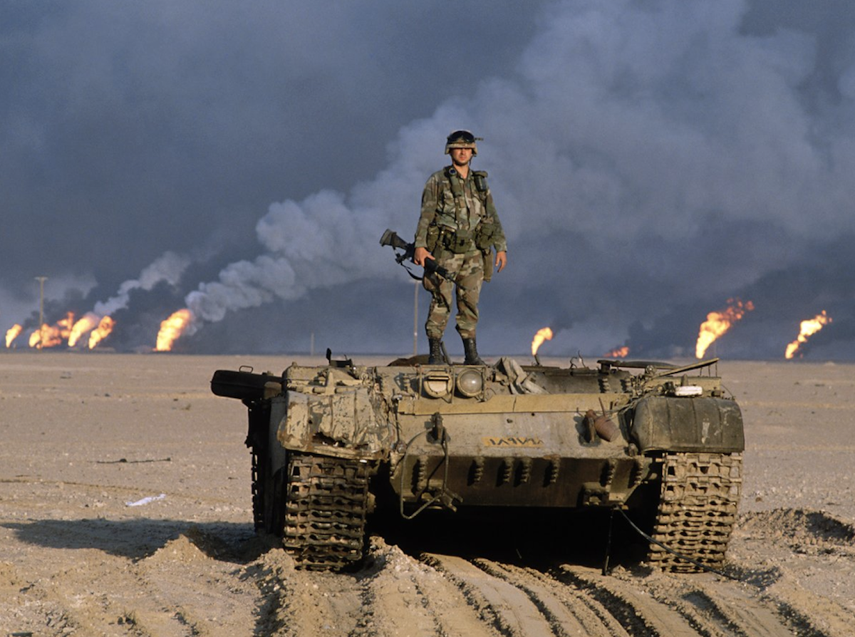 A US soldier stands atop of a destroyed Iraqi T-55 while the oil fields of Kuwait burn behind him, set ablaze by retreating Ba'athist forces during the 1st Gulf War; northern Kuwait, early 1991